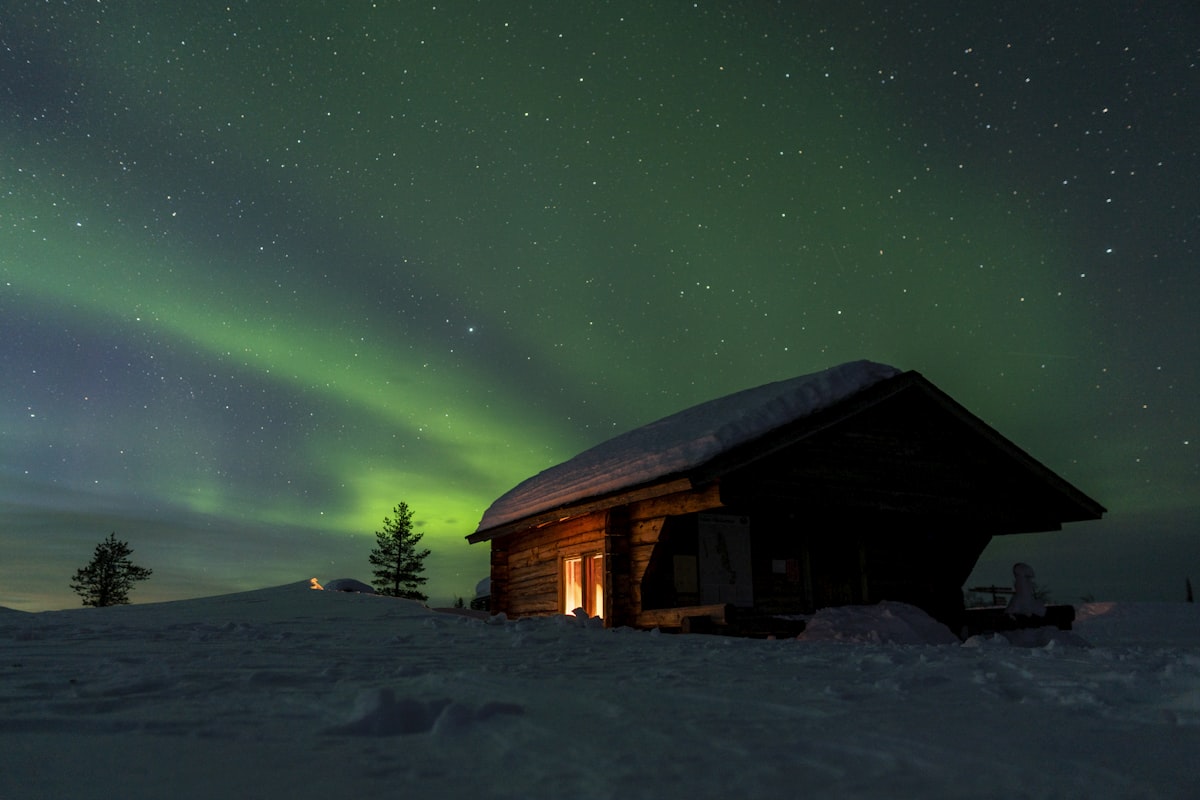 A small cabin with a warmly lit doorway standing in deep snow under a green aurora and a dense field of stars — the whole argument for a dark-sky stay in one picture.