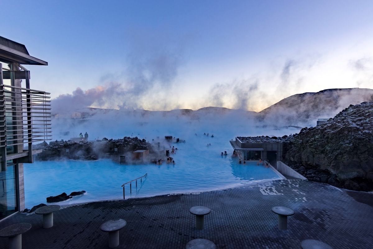 Steam drifts across the milky blue geothermal water of Iceland's Blue Lagoon, with dark lava rock around the edge of the pool and low light in the sky behind.