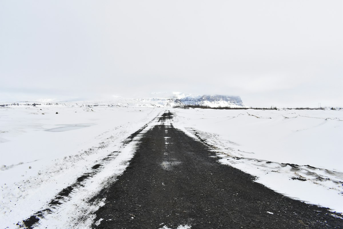 A dark gravel road running straight across a snow-covered plain in Iceland towards a flat-topped mountain under a white winter sky.