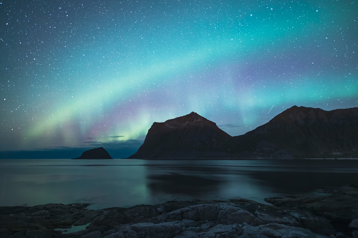 The aurora arcing over snow-capped peaks and dark water at Lofoten in northern Norway, the kind of Arctic coastline travellers weigh against Iceland when choosing a northern lights trip.