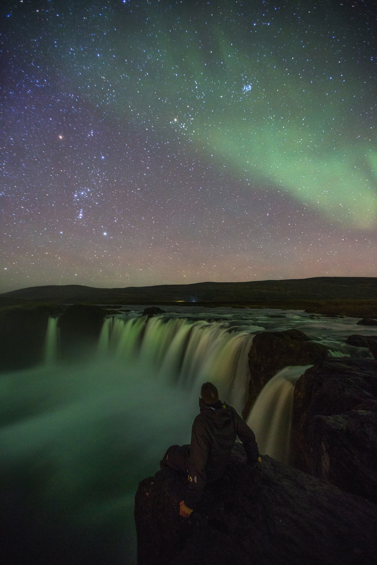A lone figure sitting beside an Icelandic waterfall at night watching green aurora borealis ripple across the sky, the kind of scene the Golden Circle offers after dark.
