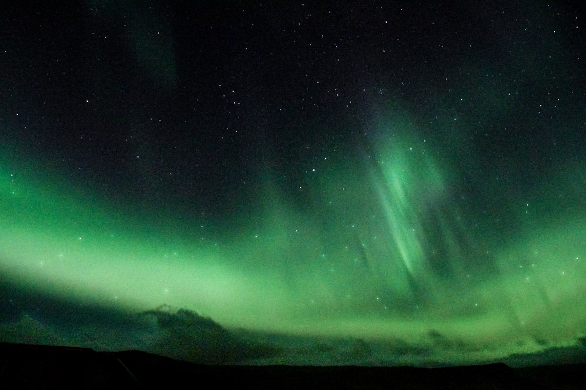 Green aurora arcing over a dark Icelandic field under a starry sky — the kind of faint first display that returns to Iceland in the final week of August.