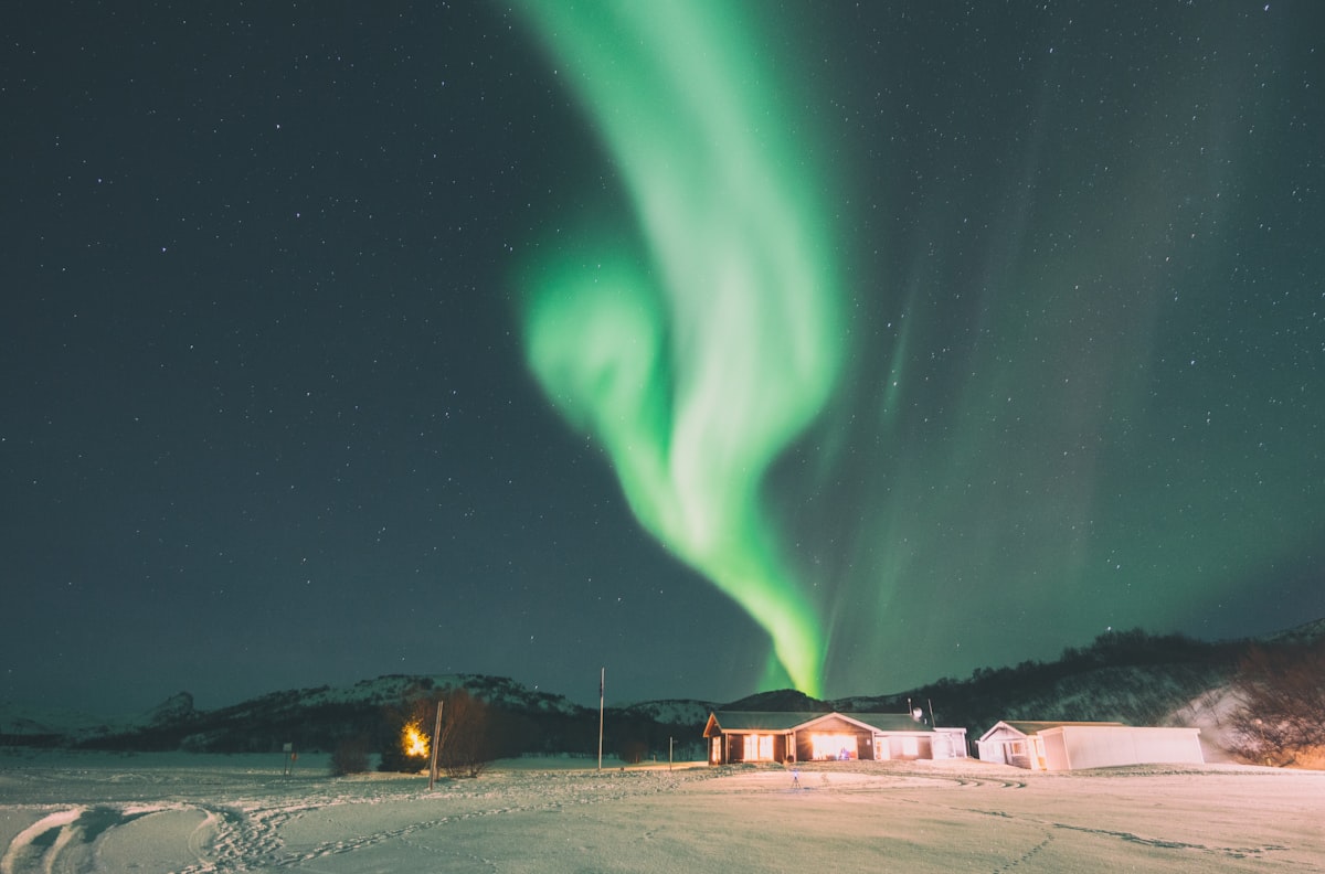 A lone cabin sitting in deep snow beneath a vivid green aurora filling the Icelandic night sky — the kind of long, dark December night that defines Iceland's midwinter.