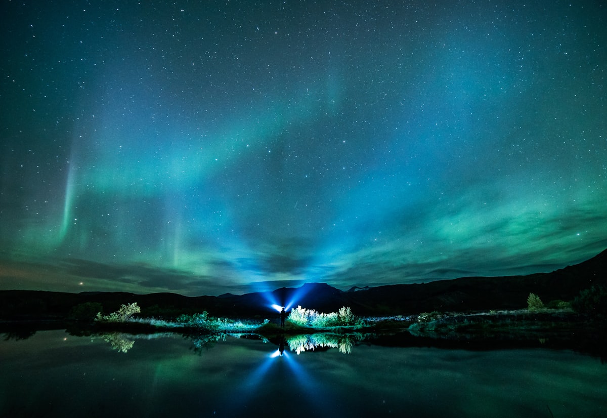 Green aurora arcing over still water under a star-filled Iceland sky — the long, dark, reflective nights that October brings back to the aurora calendar.