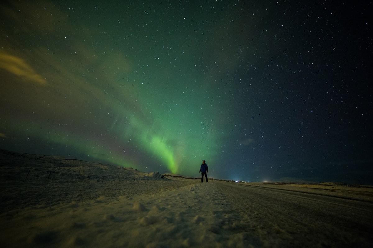 A solitary silhouette standing under bright bands of green aurora in Iceland — the exact situation that demands gear built for standing still in the cold.