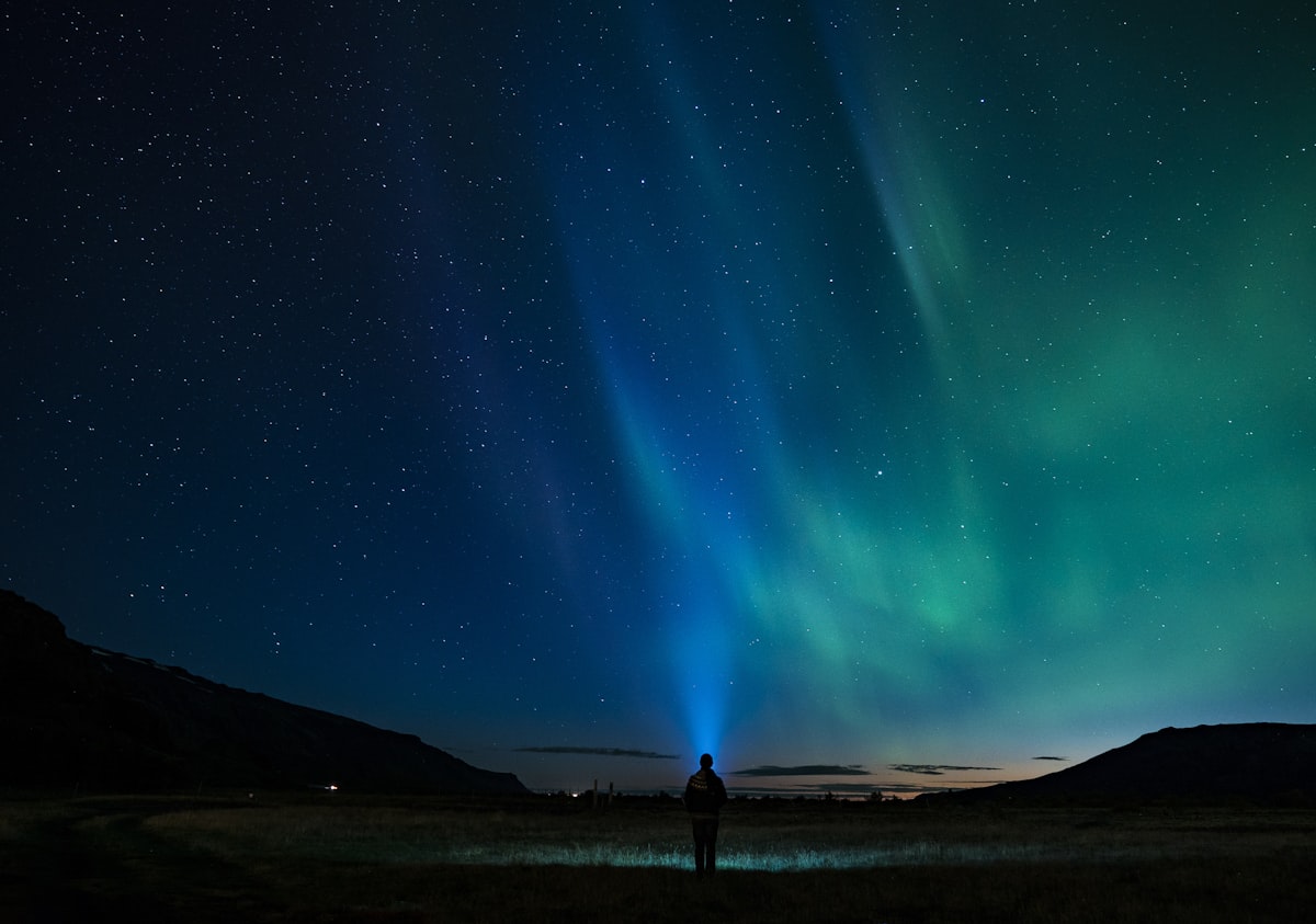 The silhouette of a lone person standing beneath a sweeping green aurora that fills the night sky over Iceland.