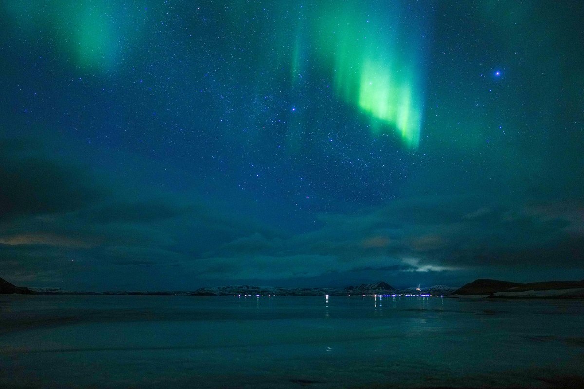A green aurora hanging over Lake Mývatn at night, with the scattered lights of a settlement along the far shore