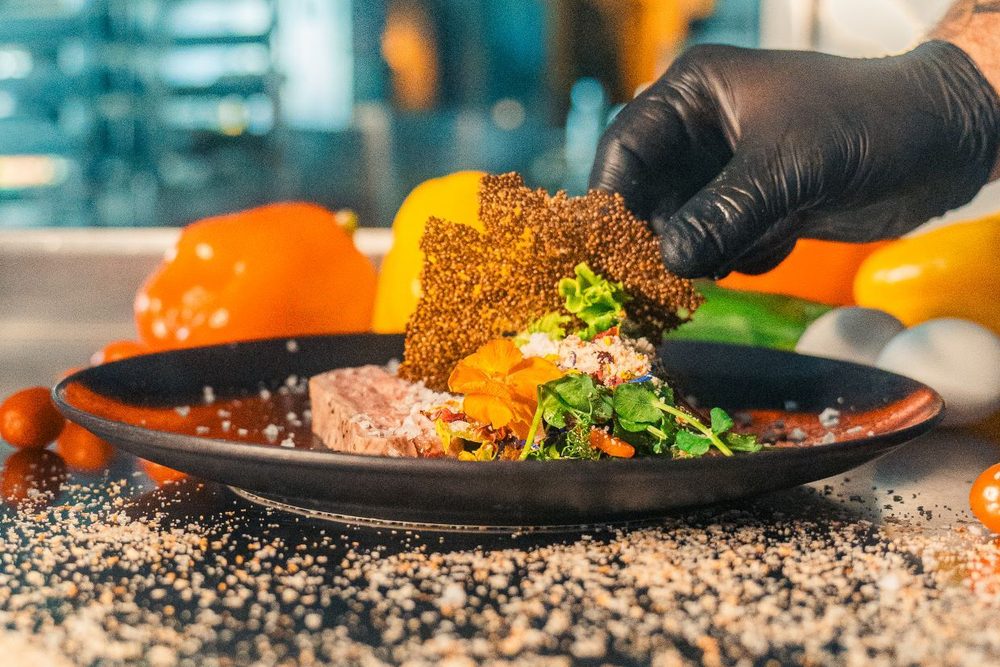 A chef in a black glove finishing a plate at Landhotel, laying a seed tuile over sliced meat, an edible flower and microgreens